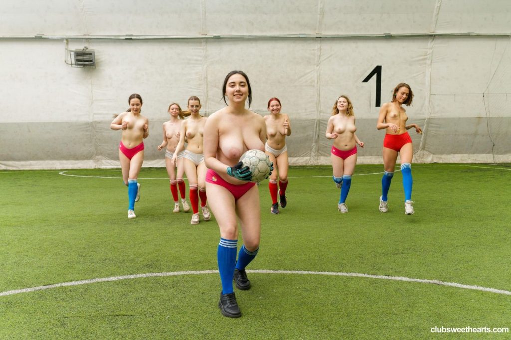 Far away view of a group of topless women Soccer players running towards the camera on an indoor pitch. The woman in the front is holding a Soccer ball in one of her hands.