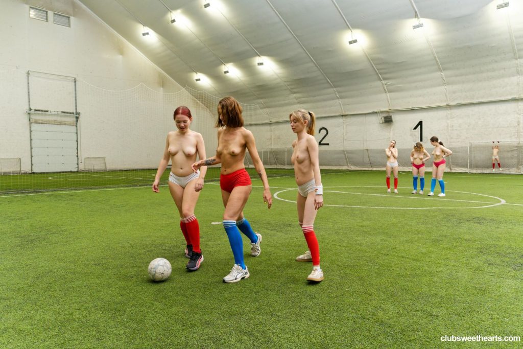Three topless Soccer players stand behind a ball on an indoor pitch. In the background three other topless players are standing.
