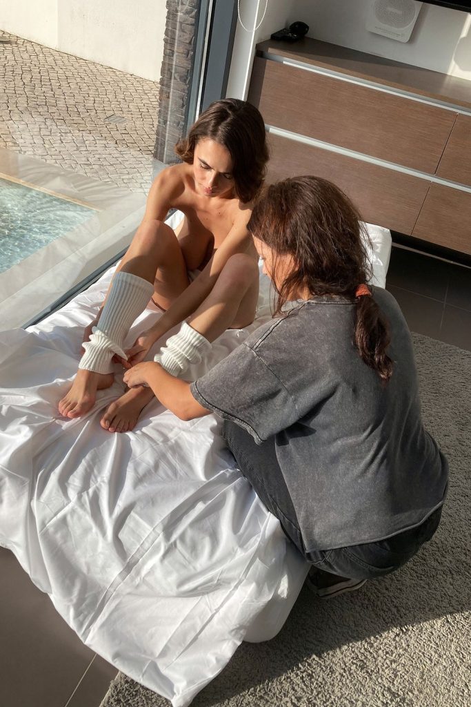 A woman sits on a bed near a large window while another person adjusts her leg warmers. Sunlight streams through the window, illuminating the scene. A swimming pool and outdoor tiles are visible outside.
