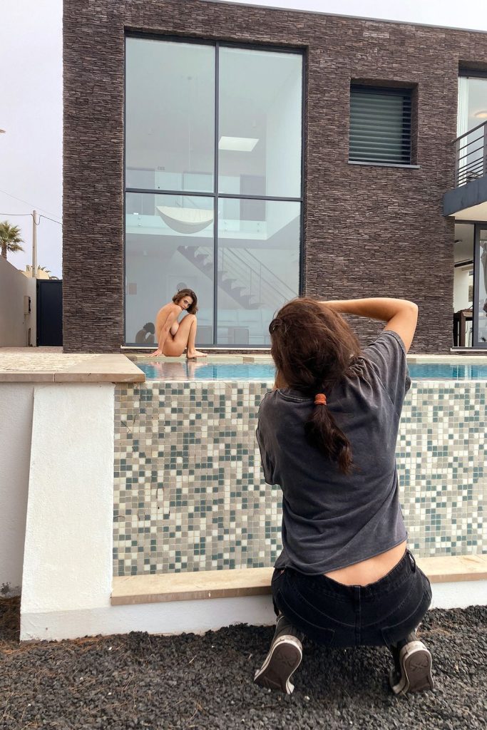 A photographer kneels by an empty pool, taking pictures of a person posing on the edge in front of a modern, glass-fronted building.
