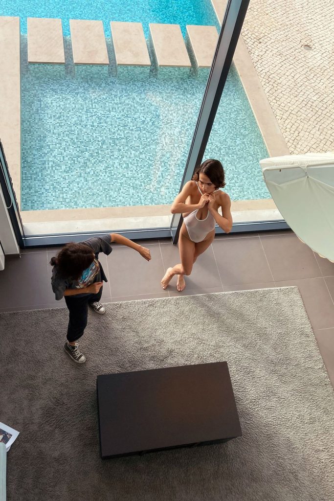View from above of two people beside a large window overlooking a swimming pool; one person is in a swimsuit and barefoot, the other is dressed casually in sneakers. They stand on a rug in a modern indoor space.