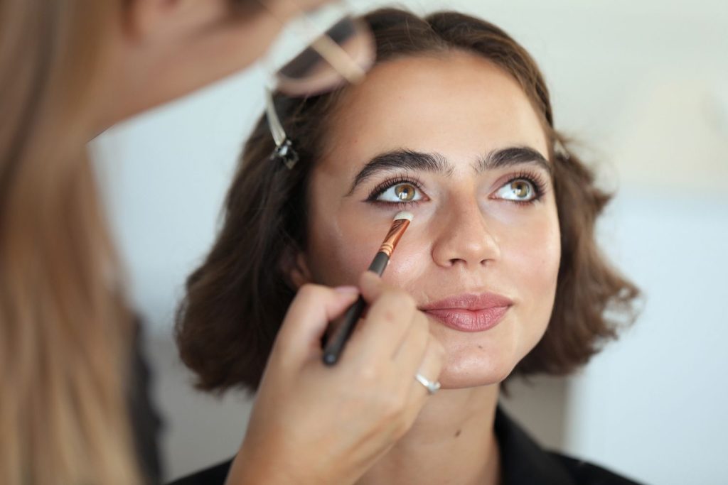 A person applies makeup with a brush under the eye of a woman with short brown hair and bold eyebrows, who is looking upwards and smiling slightly.
