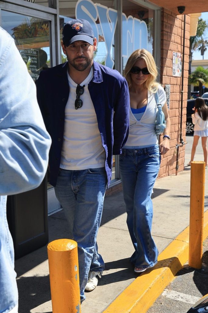 A man in a blue jacket and cap walks alongside a woman in sunglasses, a white top, and blue jeans on a sunny sidewalk outside a storefront. Yellow parking bollards are visible in the foreground.