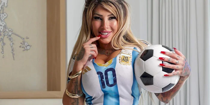 A woman with long blond hair wearing an Argentina soccer jersey holds a soccer ball and smiles, touching her lips with one finger. She has tattoos, long red nails, and is standing indoors near a window with curtains.