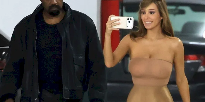 A man in a black jacket walks beside a woman in a strapless beige dress who is taking a selfie with her phone in what appears to be a parking garage.