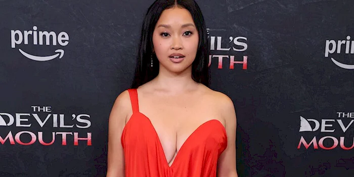 Close up of Asian woman wearing a low cut red dress in front of a dark backdrop with logos.