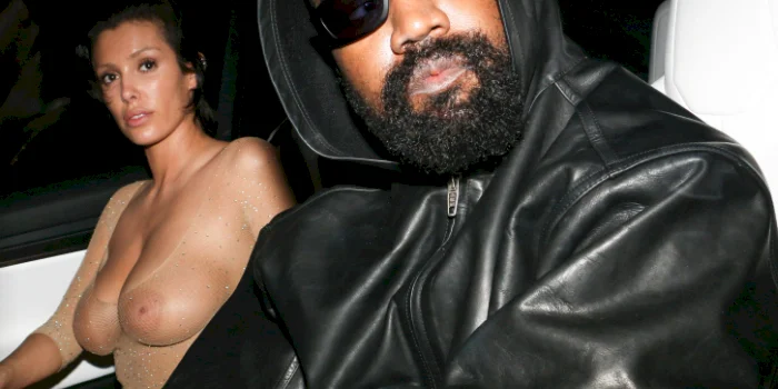 Close up of a man in a black leather jacket and sunglasses sits in a car next to a woman wearing a sheer outfit. Both are looking toward the camera.