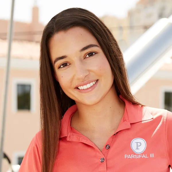 A woman with straight brown hair smiles at the camera, wearing a coral Parsifal III polo shirt, standing outdoors with blurred buildings and structures in the background.