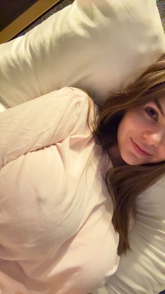 A girl with long brown hair lies on a bed, wearing a light pink shirt, resting their head on a white pillow and smiling slightly at the camera.