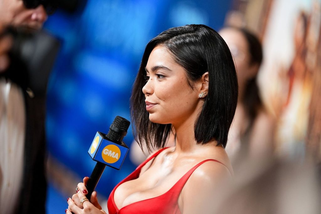 A woman in a red dress holds a GMA microphone while being interviewed at an event, with a blurred blue and gold background behind her.