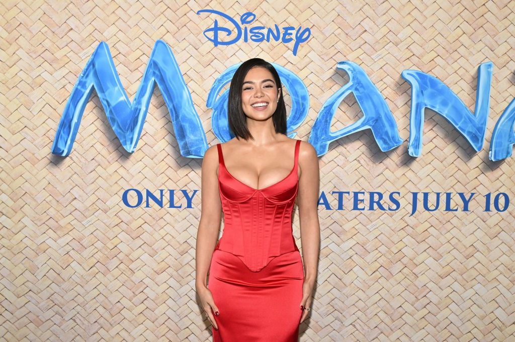 A woman in a red dress smiles in front of a Disney Moana promotional backdrop with the text ONLY IN THEATERS JULY 10.
