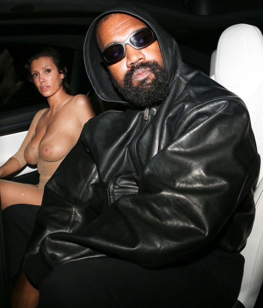 A man in a black leather jacket and sunglasses sits in a car next to a woman wearing a sheer, nude-colored outfit. Both are looking toward the camera 1 / 4.