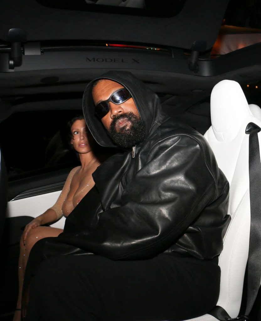 A man in a black leather jacket and sunglasses sits in a car next to a woman wearing a sheer, nude-colored outfit. Both are looking toward the camera 2 / 4.