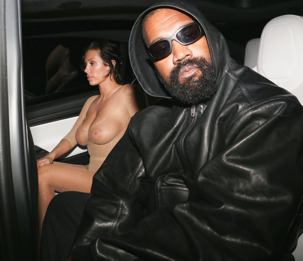 A man in a black leather jacket and sunglasses sits in a car next to a woman wearing a sheer, nude-colored outfit. Both are looking toward the camera 4 / 4.