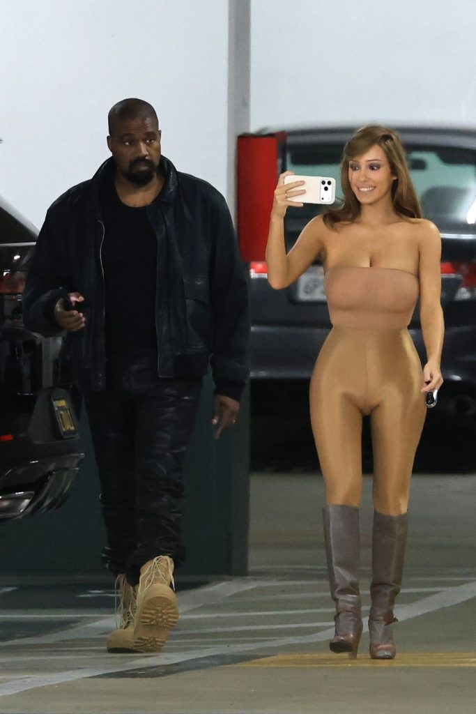 A man in dark clothing walks next to a woman wearing a nude-colored outfit and boots, who is taking a selfie in a parking garage.