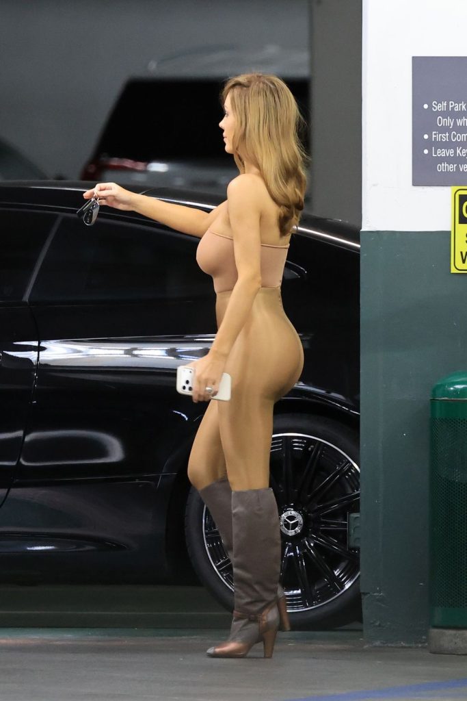 A woman in a form-fitting nude outfit and heeled boots walks in a parking garage, holding sunglasses and a phone, with cars and a green trash can nearby.