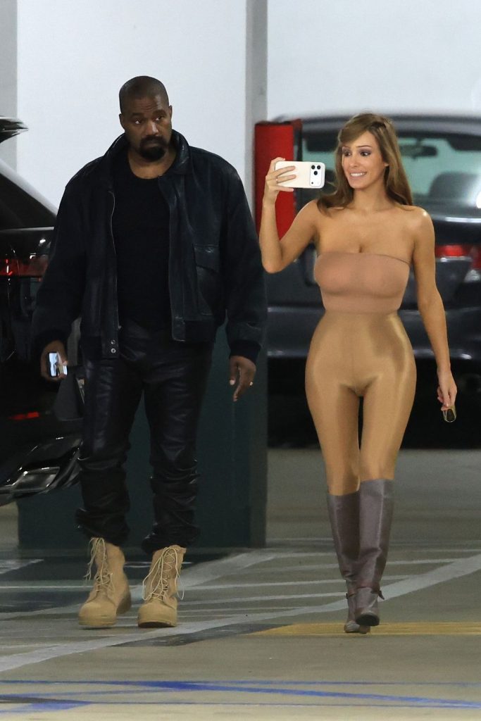 A man in a black jacket and boots stands next to a woman in a nude bodysuit and gray boots taking a selfie in a parking garage. Cars are parked in the background.