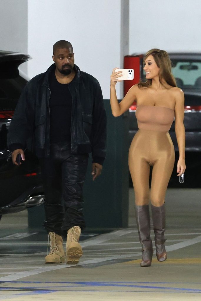 A man in dark clothes walks beside a woman in a nude-colored, form-fitting outfit and boots, who is taking a selfie with her phone in a parking garage.