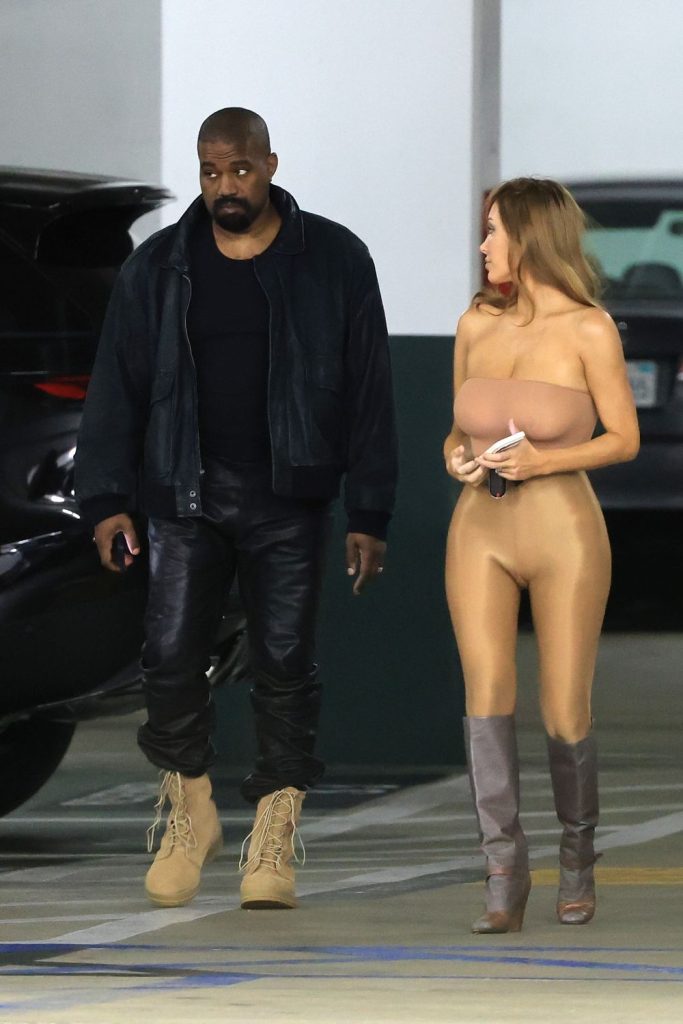 A man in dark clothing and tan boots walks beside a woman wearing a nude-toned, form-fitting bodysuit and tall boots in a parking garage.