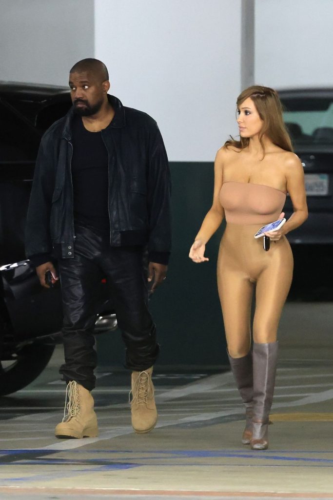 A man in a black jacket and tan boots walks beside a woman in a fitted nude-colored outfit and gray boots in a parking garage.