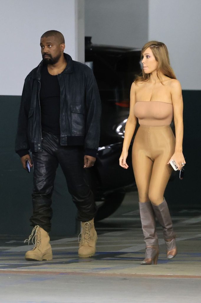 A man in dark leather clothes and tan boots walks beside a woman in a nude-colored bodysuit and heeled boots in a parking garage. Both hold phones and look ahead.