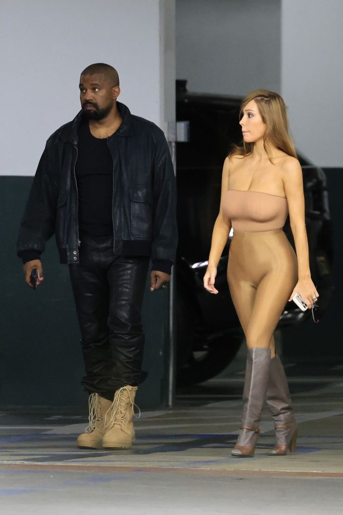 A man in dark clothing and boots walks beside a woman wearing a nude-colored, form-fitting outfit and gray boots in a parking garage.