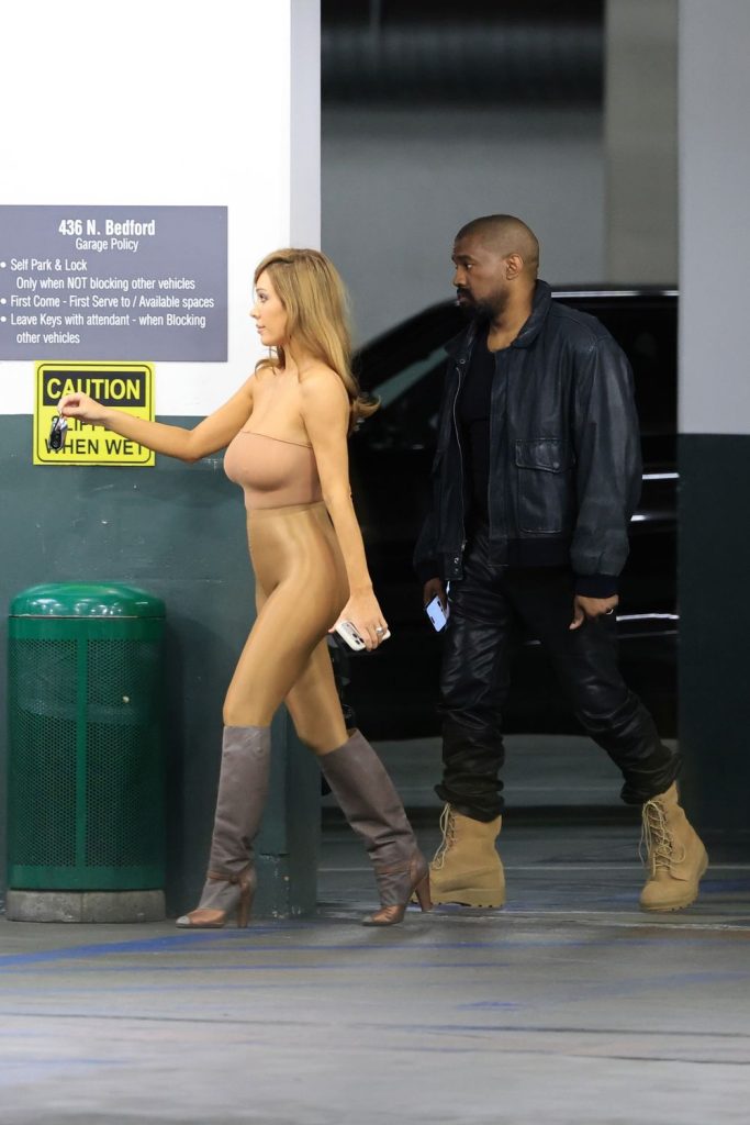 A woman in a revealing outfit and boots walks beside a man in dark clothing and tan boots in a parking garage. There is a caution sign and a green trash bin nearby.