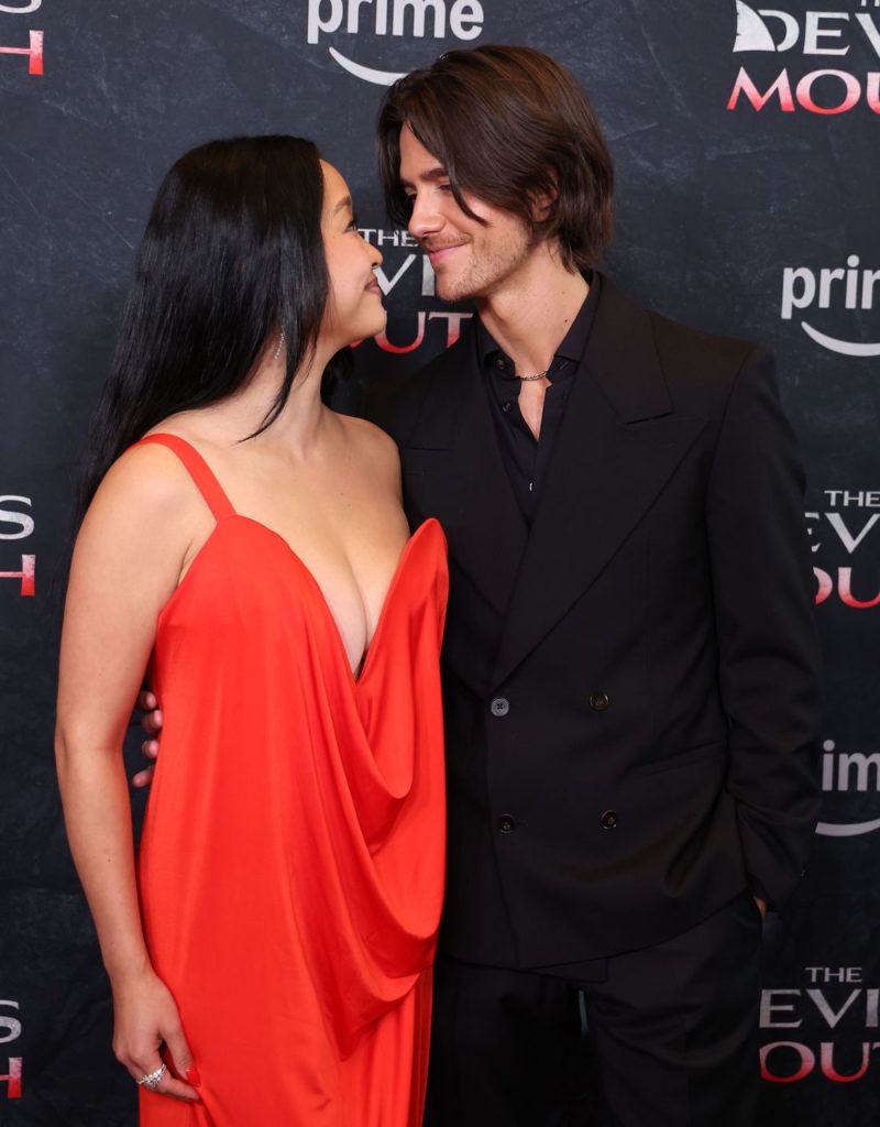 A woman in a red dress and a man in a black suit smile at each other on a red carpet with a backdrop featuring prime and THE DEVILS MOUTH logos.