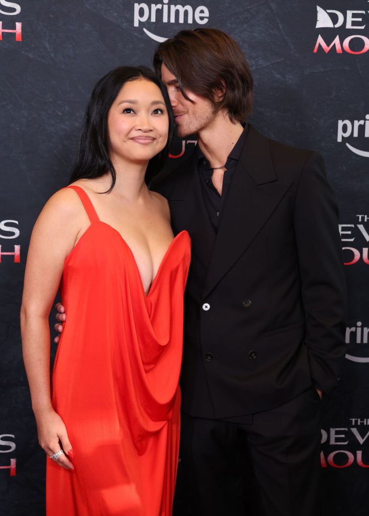Two people pose on a red carpet. The woman wears a red dress and smiles while the man in a black suit leans in, appearing to whisper in her ear. The background features the words The Devils Mouth and Prime..