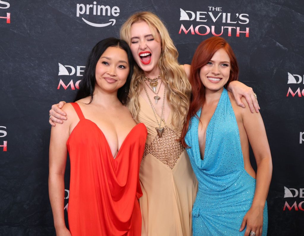Three women in elegant gowns pose together, smiling and laughing, at a red carpet event for The Devils Mouth, with a step-and-repeat banner and Prime logo in the background.