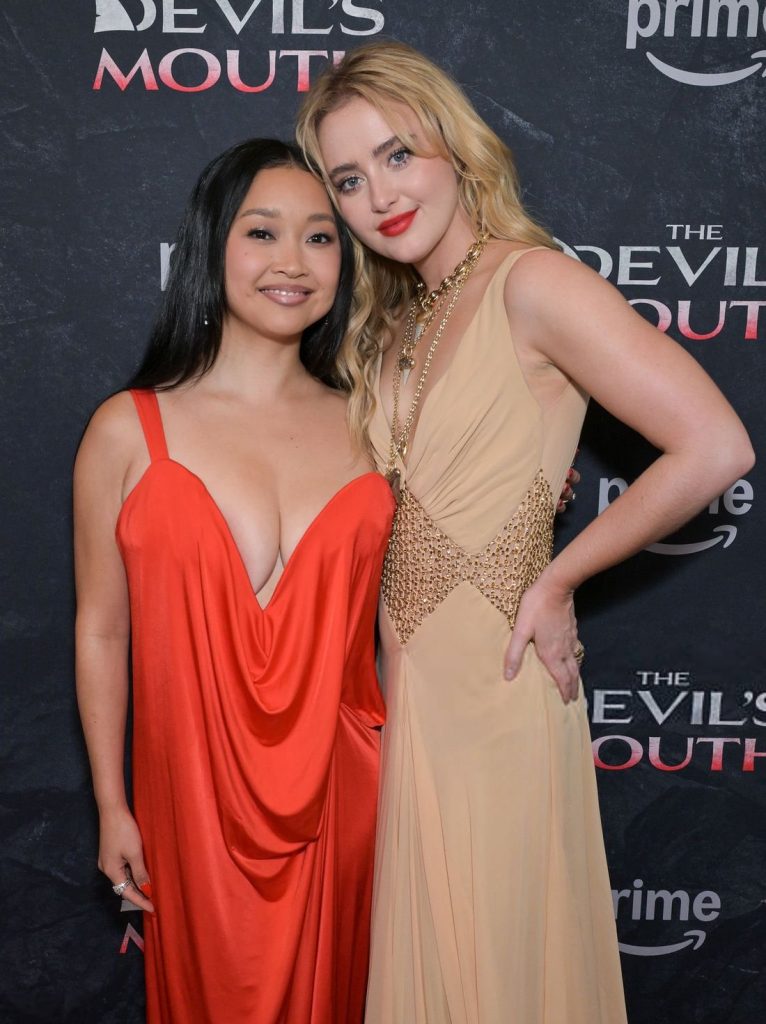 Two women in elegant dresses pose arm-in-arm and smiling on a red carpet in front of a backdrop with the words THE DEVILS MOUTH and prime video printed on it.