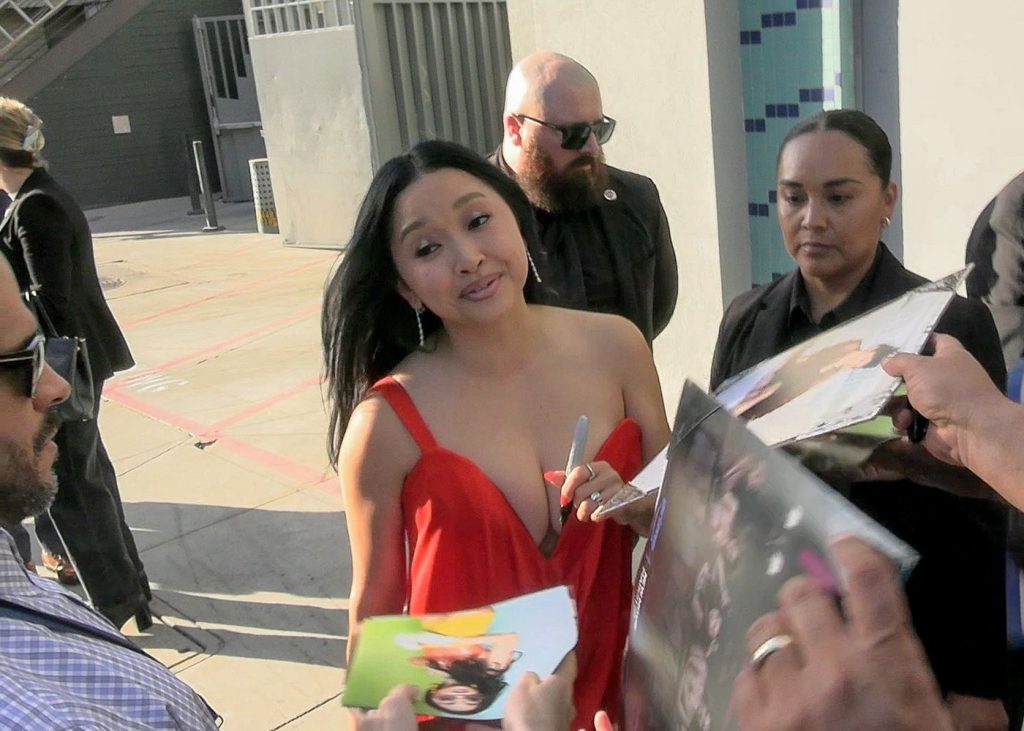 A woman in a red dress signs autographs for fans outdoors, surrounded by security guards and people holding photos and pens.