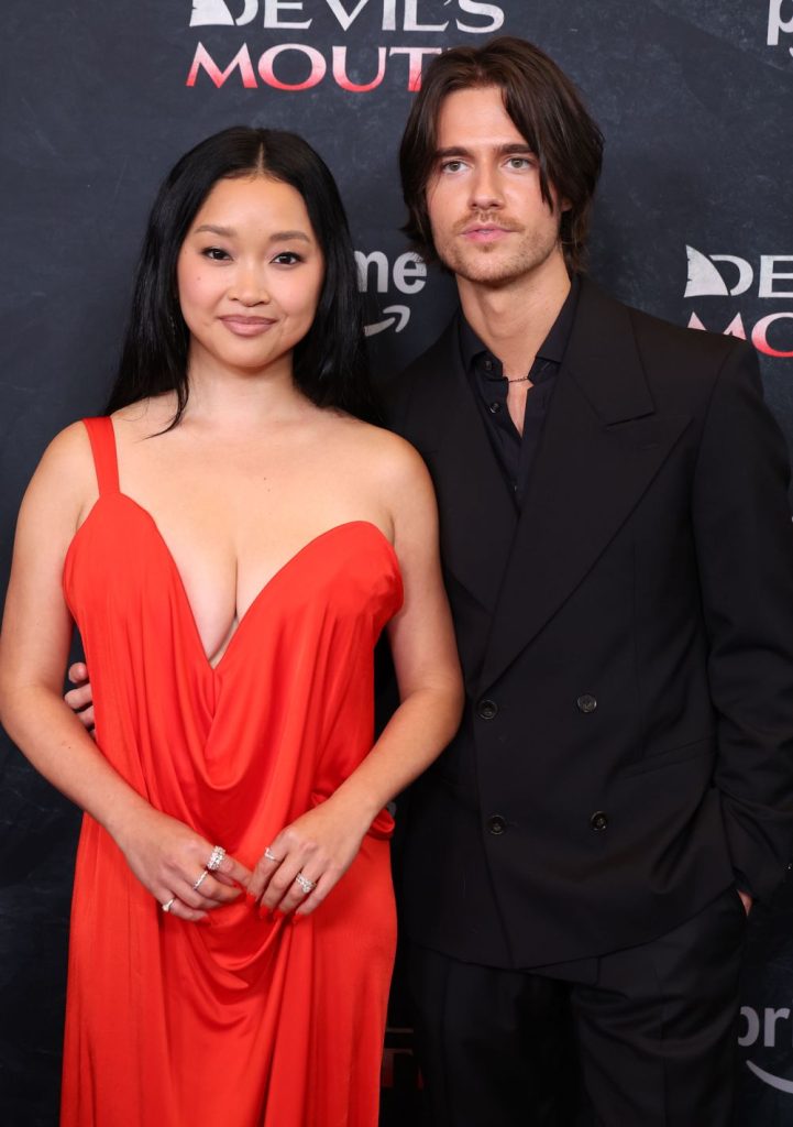 Two people posing together at an event, one wearing a bright red dress and the other in a dark suit, standing in front of a black backdrop with white and red text.