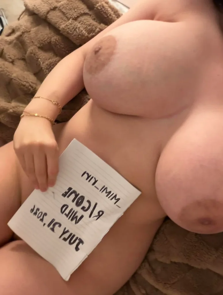 Close up body shot of a naked girl lying back and holding a paper with backwards text on it in front of her crotch.