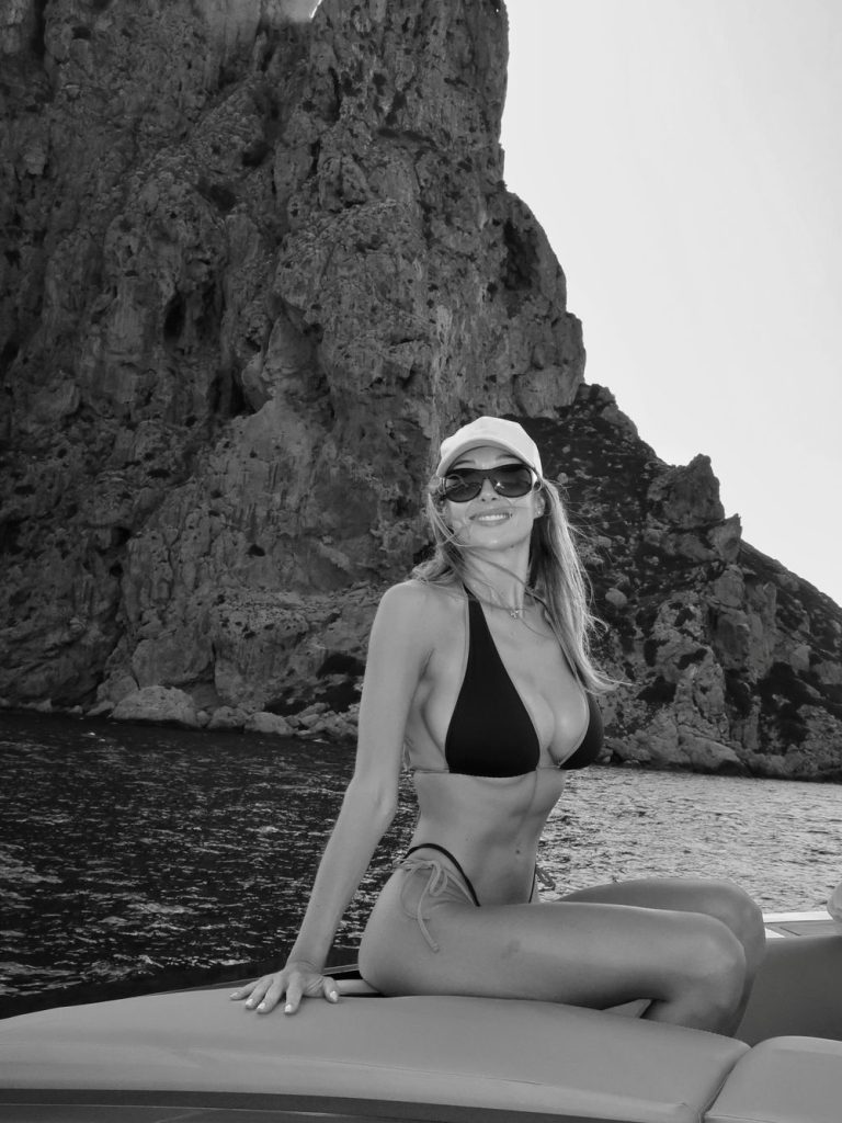 A woman in a bikini, sunglasses, and a cap sits on a boat in front of a rocky cliff by the sea. The photo is black and white.