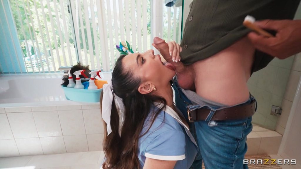 Side view of a maid kneeling in front of a man with pulled down jeans. She holds and licks the bottom of his erect penis.