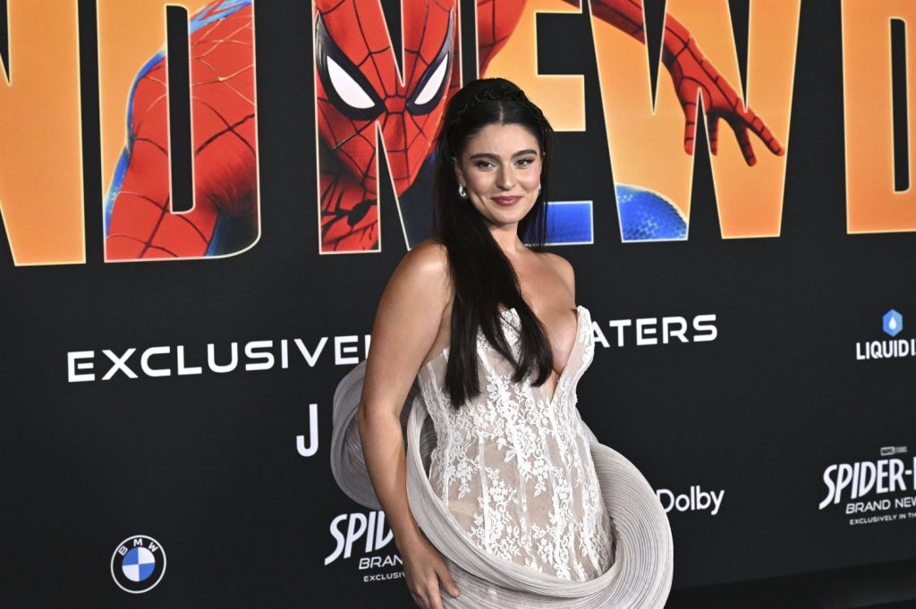 A woman in a sheer white dress poses on a red carpet in front of a Spider-Man movie backdrop featuring Spider-Mans arm and bold lettering.
