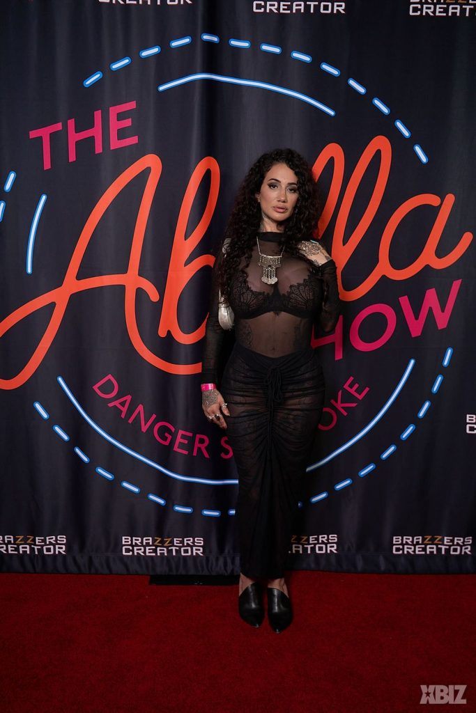 A woman with long curly hair in a sheer black dress poses on a red carpet in front of a backdrop that reads The Abella Show and Brazzers Creator.