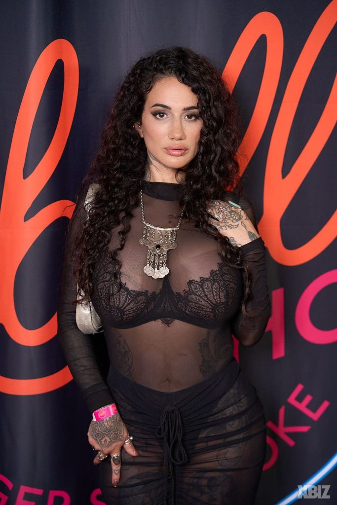 A woman with long curly hair wears a sheer black lace outfit and statement necklace, posing in front of a backdrop with large orange lettering. She has visible tattoos on her hands and arms.