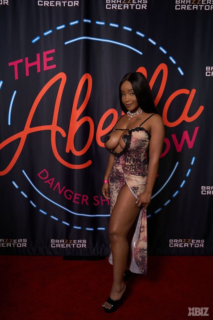 A woman in a revealing dress poses on a red carpet in front of a backdrop that reads The Abella Show, with additional logos for Brazzers Creator and XBIZ.