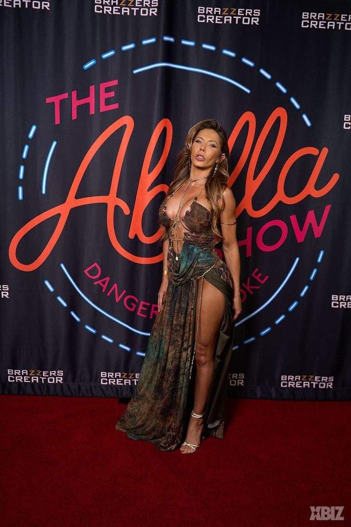 A woman in a revealing green dress poses on a red carpet in front of a backdrop reading The Abella Show with Brazzers Creator and XBIZ logos.