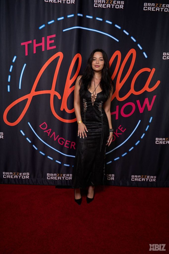 Woman in a black dress poses on a red carpet in front of a backdrop reading The Abella Show, with additional text and logos in the background.