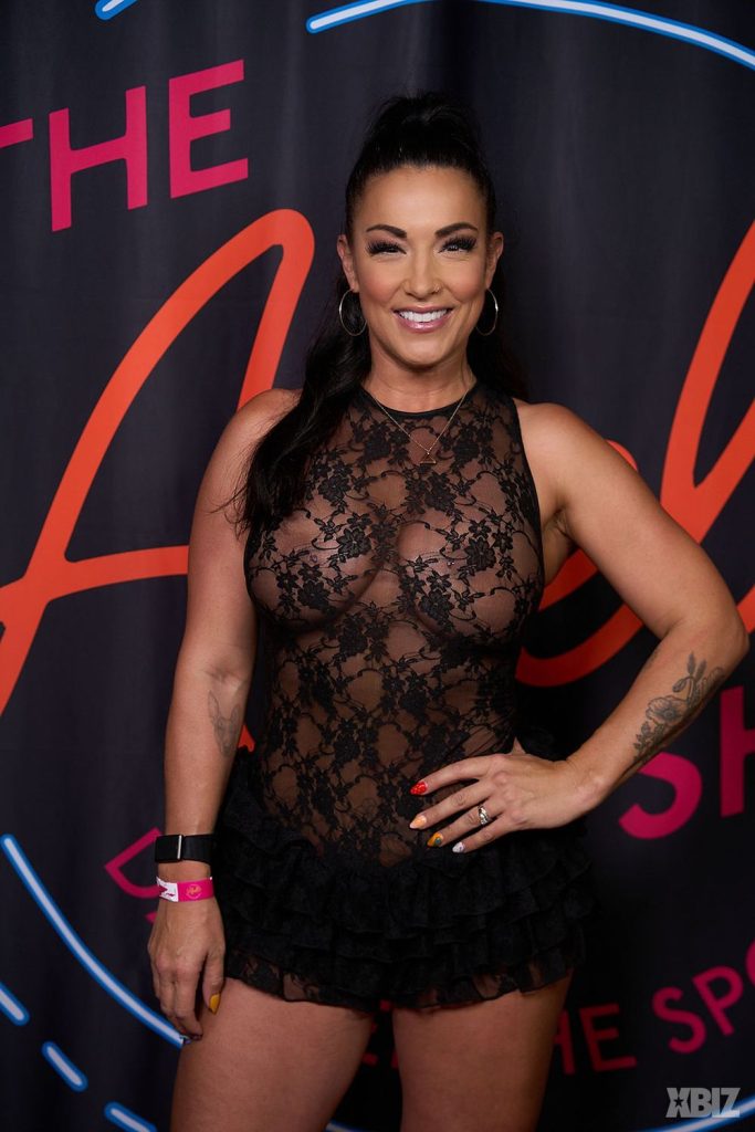 A woman in a sheer black lace dress poses, smiling, with one hand on her hip, in front of a dark backdrop with bright pink and blue writing.