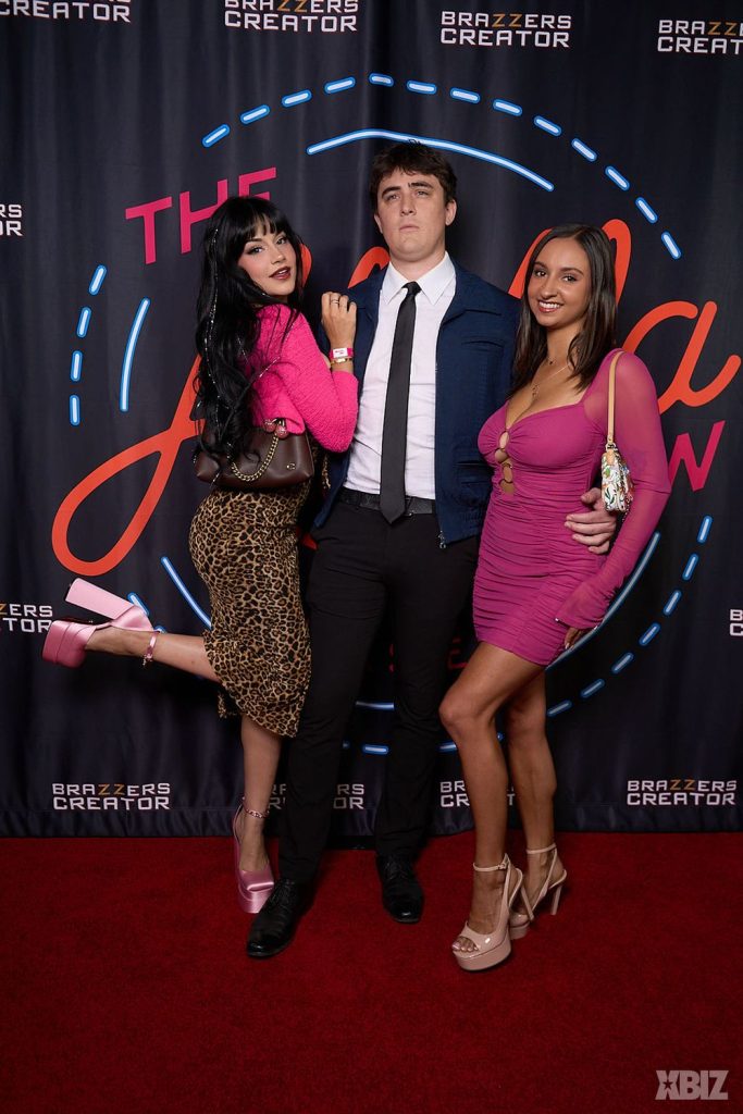 Three people pose on a red carpet in front of a Brazzers Creator backdrop. Two women in pink outfits stand on either side of a man in a blue jacket, white shirt, and tie.