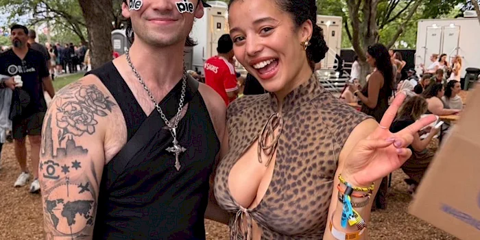 Two people pose outdoors at a festival. The man wears sunglasses with die written on them and has tattoos, while the woman smiles, wearing a leopard print outfit and giving a peace sign. Other festivalgoers are in the background.