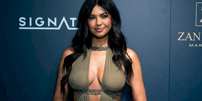 Belle Hassan wearing a sleeveless, olive-green, cut-out dress stands in front of a dark blue backdrop with white and gold logos. She is smiling slightly and has her hands in front of her.