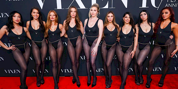 Nine women in matching black bodysuits and sheer black tights pose together on a red carpet in front of a VIXEN 10 Year Anniversary backdrop.