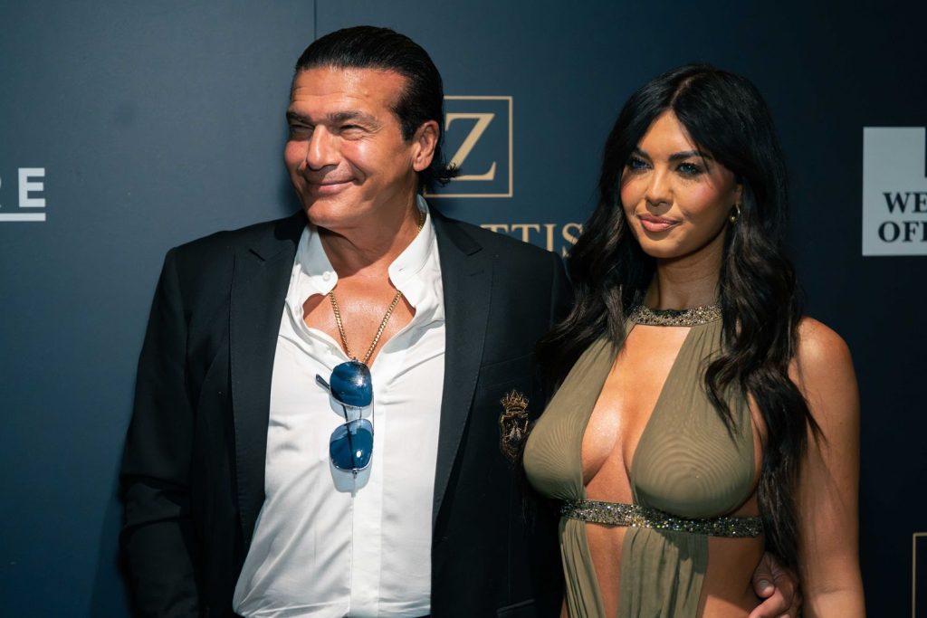 A man in a dark suit with sunglasses hanging from his shirt and a woman in a revealing dress pose together at a formal event against a dark backdrop with logos.