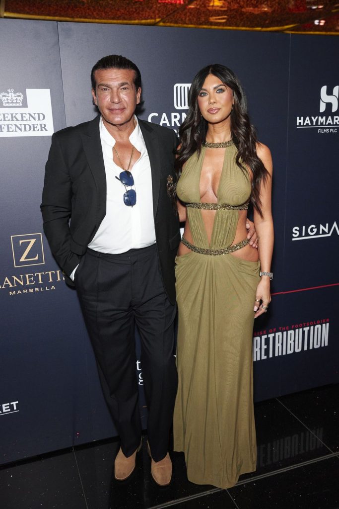 A man in a suit and a woman in a green dress pose together on a red carpet in front of a dark event backdrop with logos.