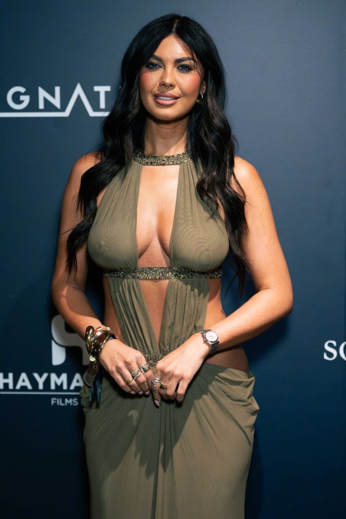 A woman with long dark hair in a sleeveless, cut-out olive dress poses in front of a dark blue backdrop with white text and logos, smiling at the camera.